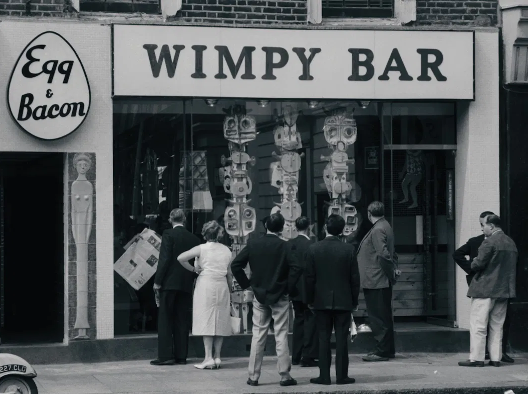 Passers-by looking at a modern sculpture in the window of a Wimpy Bar in London, c.1961. Photograph by John ‘Hoppy’ Hopkins. © Estate of John Hopkins.
