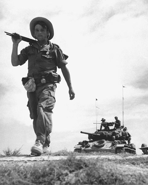 A French Foreign Legionnaire goes to war along the dry rib of a rice paddy, between Haiphong and Hanoi. Behind the Legionnaire is a U.S. gifted tank. Ca. 1954.